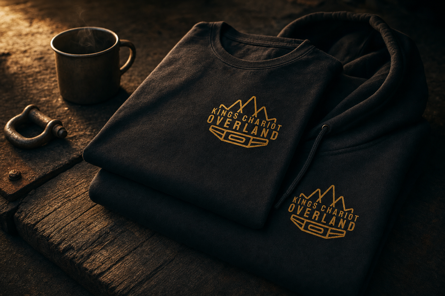 Folded heavyweight black King's Chariot Overland t-shirt and hoodie with gold logo print, photographed on dark wood