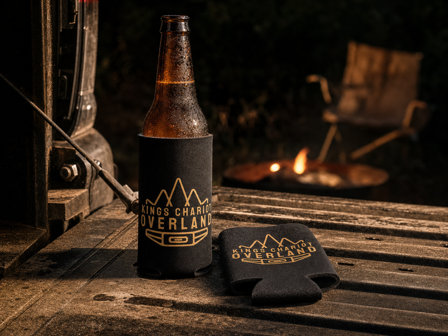 Black neoprene beer bottle insulator (koozie) with King's Chariot Overland logo screen-printed in gold, wrapped around a frosty amber bottle on a truck tailgate